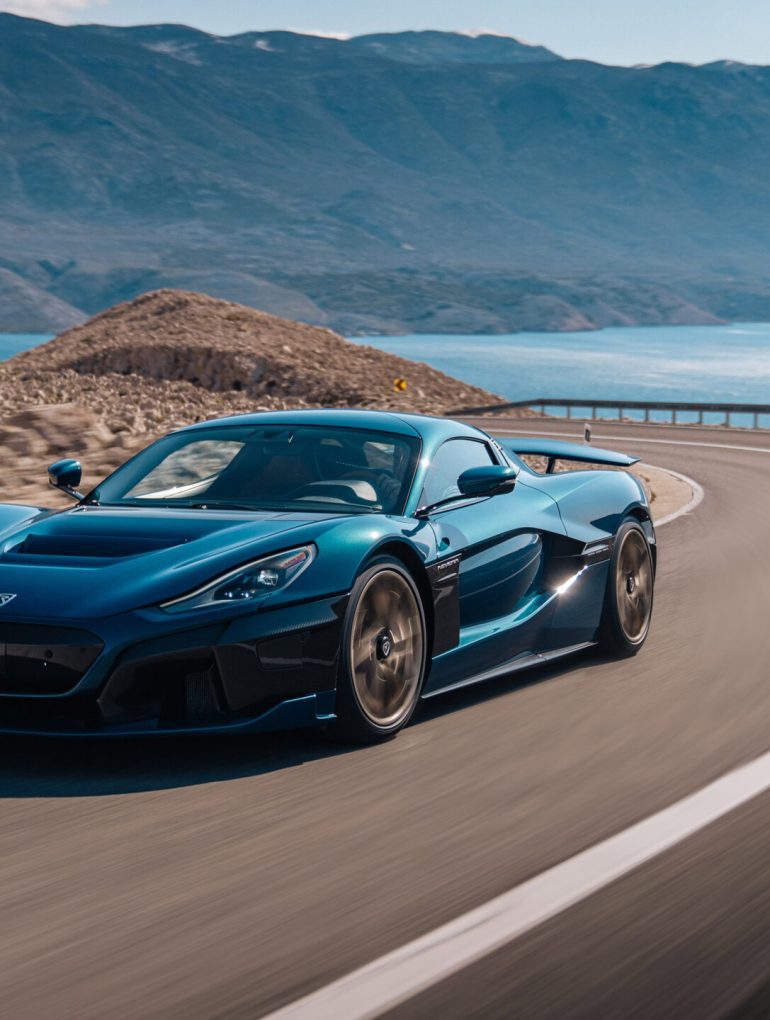 Navy blue Rimac Nevera speeding around curve on highway near water